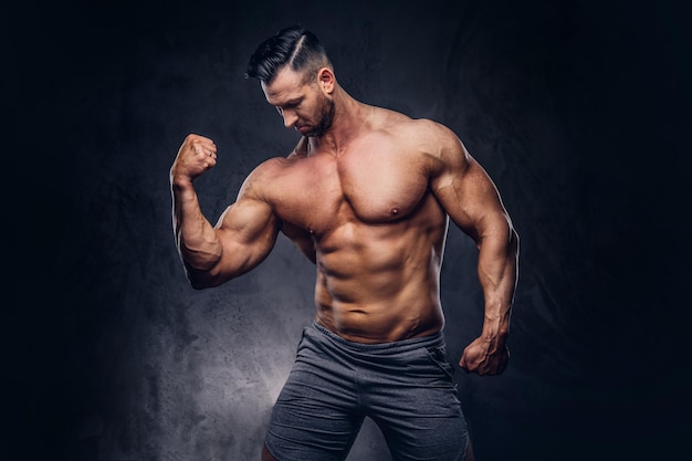 Portrait of a shirtless tall huge male with a muscular body with a stylish haircut and beard, in a sports shorts, posing in a studio. Isolated on a dark background.