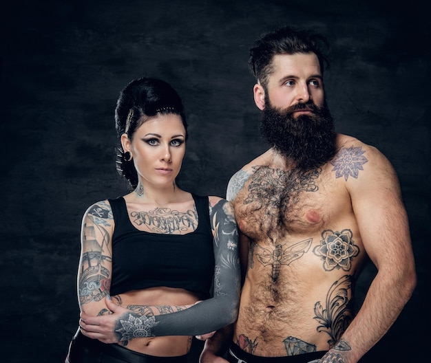 Free photo portrait of shirtless, tattooed bearded hipster male and brunette female with tattoo ink on her torso over dark grey background.