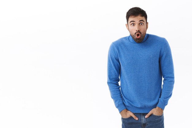 Portrait of shocked and astonished, startled handsome man with beard, popping eyes, open mouth speechless staring at something shocking, standing white wall