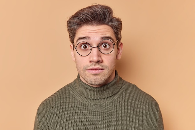 Portrait of shocked brunet European man stares surprisingly at camera keeps eyes widely opened wears round spectacles and sweater sees something breathtaking isolated over beige studio background