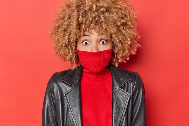 Free Photo portrait of shocked european woman stares bugged eyes covers mouth with collar of jumper wears leather jacket isolated over vivid red background reacts on something breathtaking. omg concept