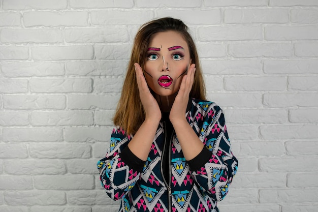 Portrait of shocked girl wearing creative makeup and holding hands to her face. High quality photo