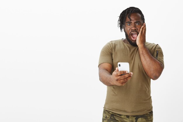 Portrait of shocked guy in a brown t-shirt posing against the white wall with his phone