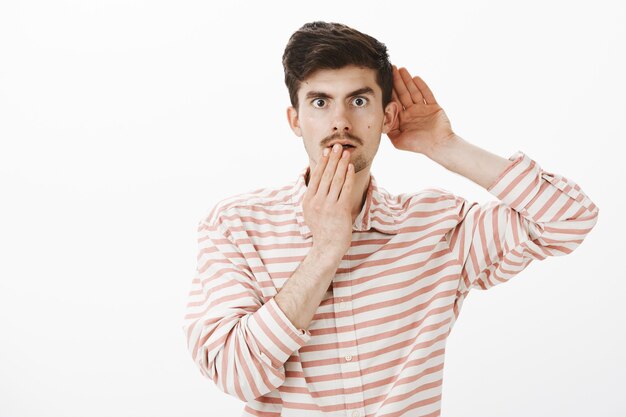 Portrait of shocked intrigued attractive caucasian man in trendy striped shirt, holding hand near ear and mouth, overhearing conversation or eavesdropping, hearing something shocking and interesting