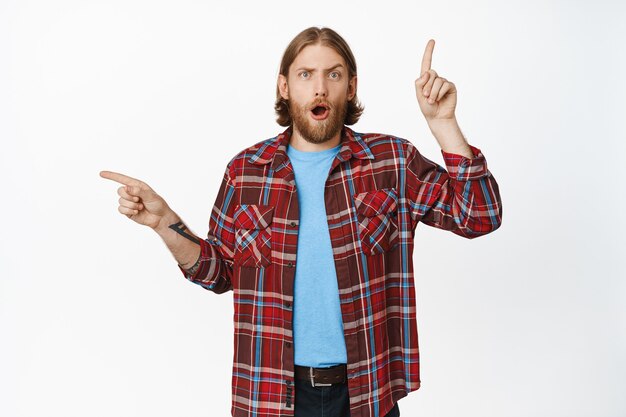 Portrait of shocked and surprised blond adult man, pointing up standing in casual clothes on white