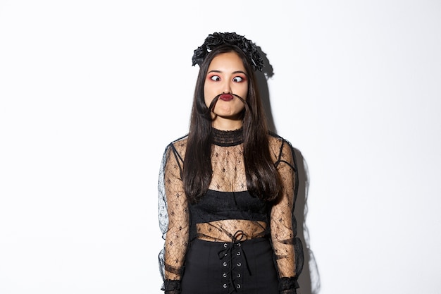 Portrait of silly and funny asian girl foolind around, grimacing and making moustache from hair strand, standing over white background in halloween costume.