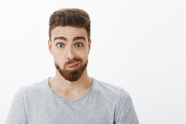 Free photo portrait of silly and funny handsome man with beard, moustache and blue eyes smirking making uncertain awkward face looking in mirror and thinking about making changes posing against gray wall