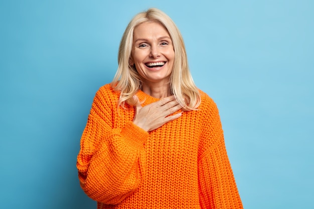 Free photo portrait of sincere blonde woman smiles broadly has white perfect teeth keeps hand on chest feels very glad to receive help from close person wears orange knitted sweater.