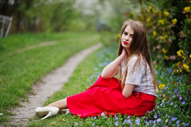 Portrait of sitiing beautiful girl with red lips at spring blossom garden on grass with flowers wear on red dress and white blouse