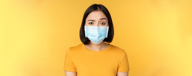 Portrait of skeptical and confused asian woman in medical face mask raising eyebrow doubtful standin