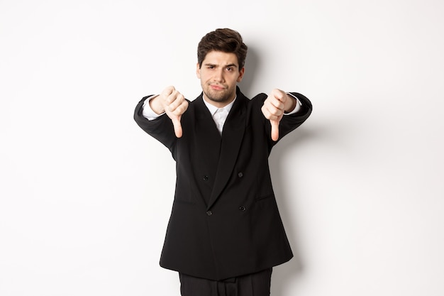 Free photo portrait of skeptical and disappointed man in black suit, frowning upset, showing thumbs-down, dislike something bad, standing over white background.