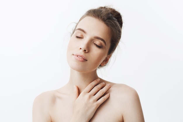 Free photo portrait of sleepy good-looking young student girl with dark hair in bun hairstyle being naked, touching shoulders with hand and closed eyes, taking shower after long day at university.