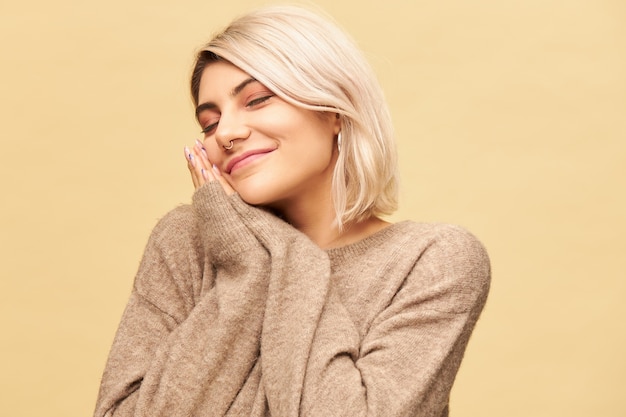 Portrait of sleepy tired young blonde woman with nose ring placing head on hands pressed together and keeping eyes closed, having nap or sleeping, smiling happily. Sleep, bedding and fatigue concept