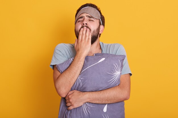 Portrait of sleepy young man with his pillow in hands, yawning and covering his mouth with palms, posing with blindfold on forehead