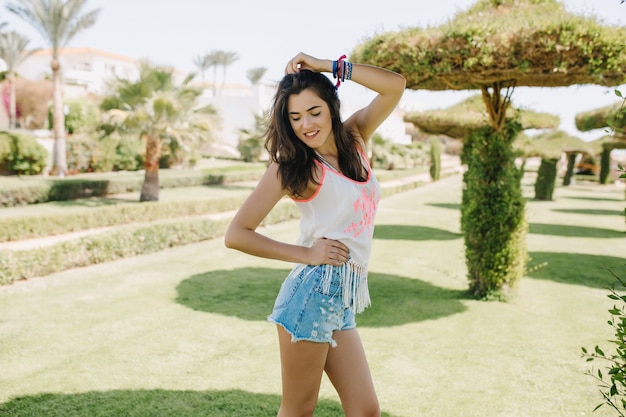 Portrait of slim charming brunette girl in denim shorts posing in exotic park in sunny day. Cheerful smiling young woman with dark hair having fun outside standing on nature