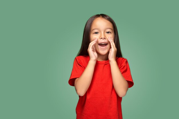 Portrait of a small pretty girl standing sideways and calling someone holding her hand near her mouth wearing a red t-shirt, green background