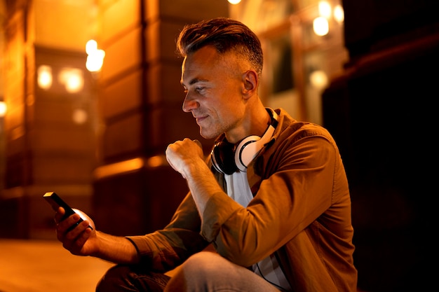 Free photo portrait of smiley man in the city at night with headphones and smartphone