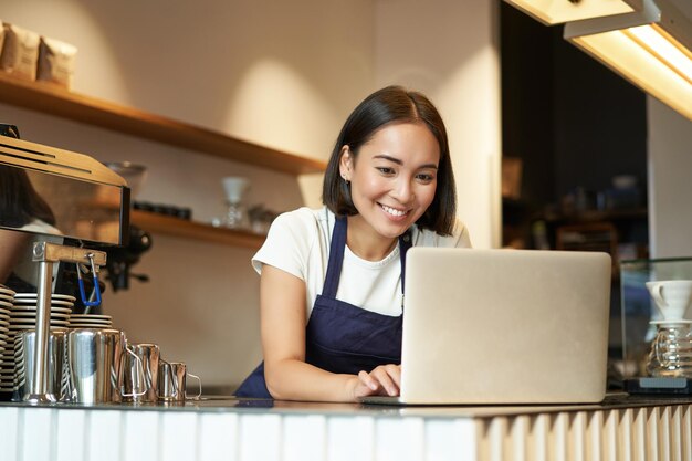 Portrait of smiling asian barista cafe owner entrepreneur working on laptop processing orders on com