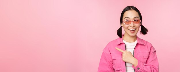 Portrait of smiling asian girl in stylish outfit sunglasses pointing finger left showing advertiseme