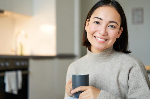 Portrait of smiling asian girl with cup of coffee drinking hot tea and getting cozy at home warming