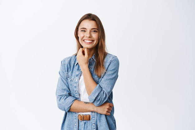 Portrait of smiling beautiful woman touching her face with natural make up and looking cheerful at front, standing against white wall