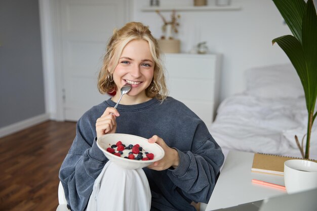 Portrait of smiling blond woman eating breakfast holding bowl and spoon sitting in bedroom looking