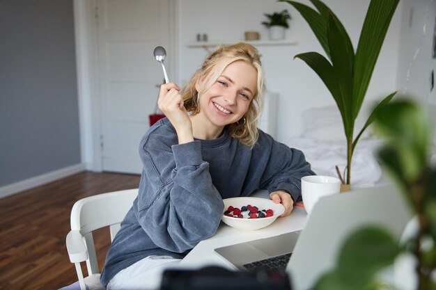 Portrait of smiling blond young woman eating in front of laptop watching videos online while having