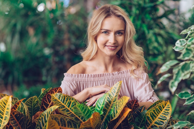 Free photo portrait of a smiling blonde young woman standing behind the garden croton leaves