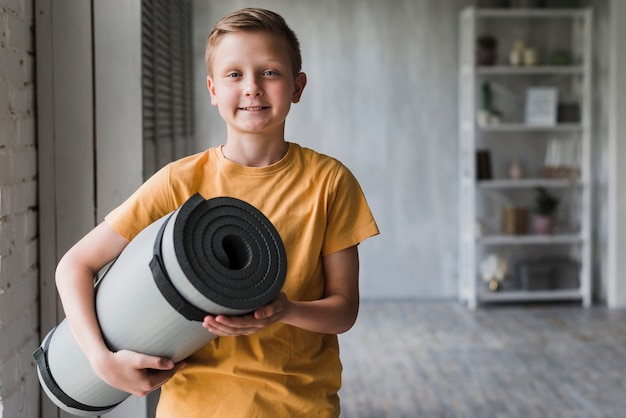 Free Photo portrait of a smiling boy holding grey rolled up exercise mat in hand