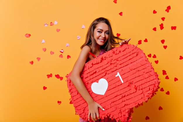 Free photo portrait of smiling brunette woman obsessed with social networks. indoor portrait of glamorous girl standing on yellow under confetti.