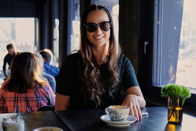 Portrait of smiling brunette woman in sunglasses, drinks morning coffee in a cafe.