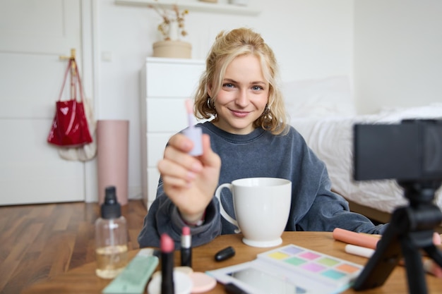 Portrait of smiling charismatic woman puts on lip gloss drinks tea records video on digital camera