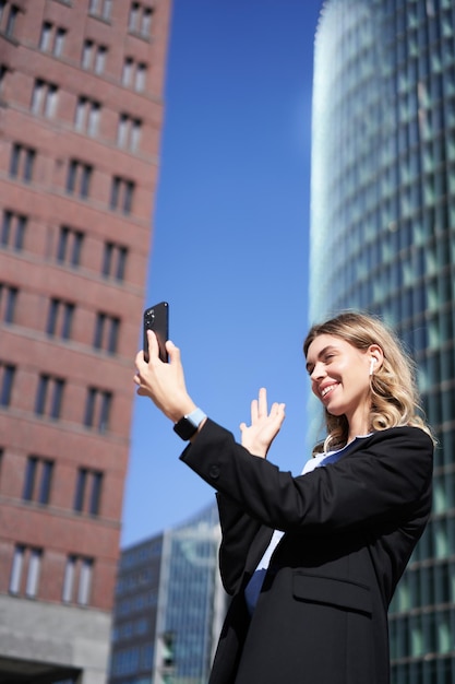 Free Photo portrait of smiling corporate woman video call on street holding mobile phone and waving at smartpho