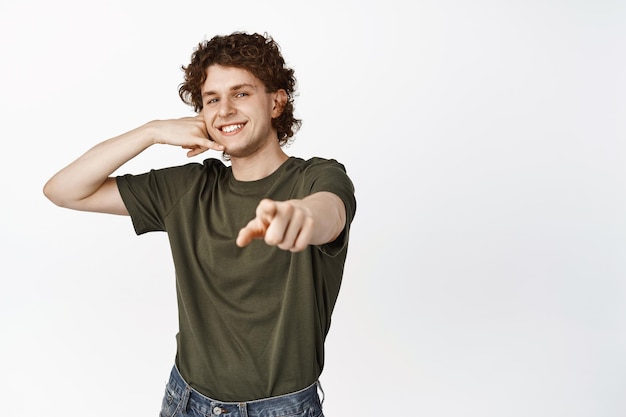 Portrait of smiling curly young man pointing finger at camera showing mobile phone gesture call me standing over white background