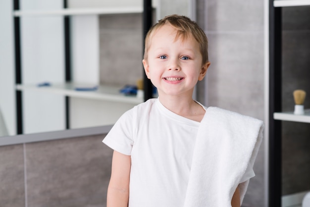 Portrait of a smiling cute little boy with white towel over his shoulder standing in the bathroom