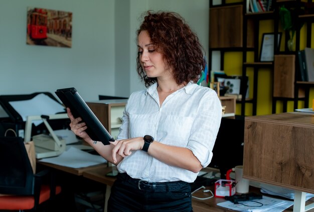 Portrait of smiling female employee holding tablet looking at it with confident smile on face standing in office