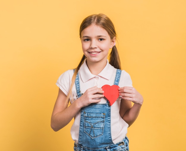 Free photo portrait of a smiling girl showing red paper cut out heart shape against yellow background