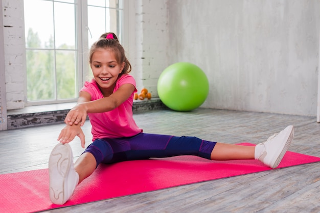 Portrait of a smiling girl sitting on exercise mat stretching his hand and leg