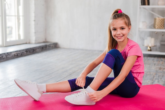 Portrait of a smiling girl sitting on exercise mat tying her shoelace