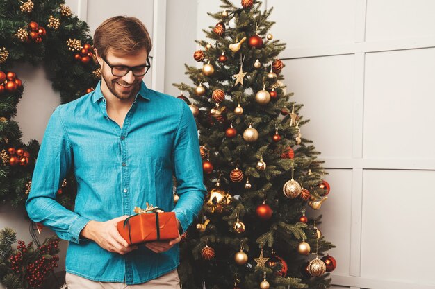 Portrait of smiling handsome man holding gift. Sexy bearded male posing near Christmas tree with present. 