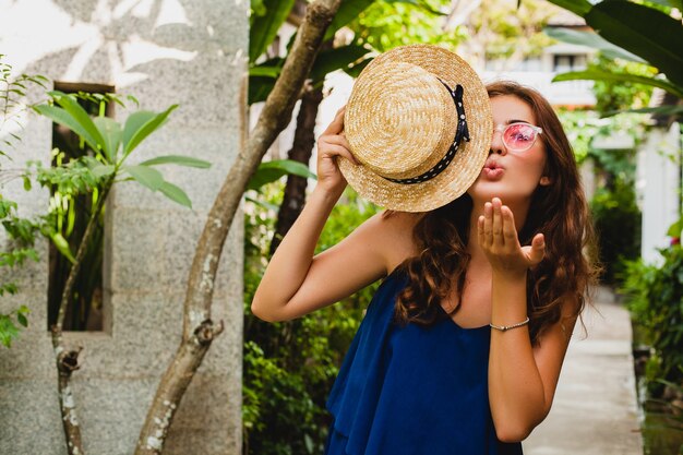 Portrait of smiling happy attractive young woman in blue dress and straw hat wearing pink sunglassses walking at tropical spa villa hotel on vacation in summer style outfit