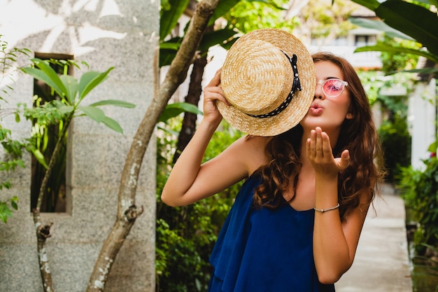 Portrait of smiling happy attractive young woman in blue dress and straw hat wearing pink sunglassses walking at tropical spa villa hotel on vacation in summer style outfit