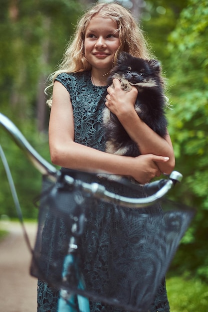 Portrait of a smiling little blonde girl in a casual dress, holds cute spitz dog. Ride on a bicycle in a park.