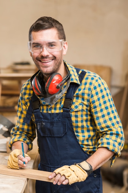 Free Photo portrait of a smiling male carpenter holding wooden plank and pencil