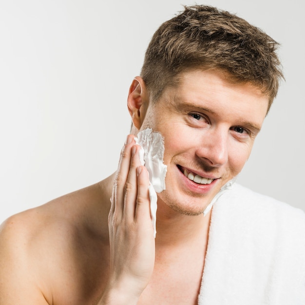 Free photo portrait of a smiling man applying shaving foam on his face with hand against white backdrop