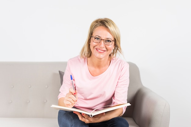 Portrait of smiling mature woman with pen and book sitting on sofa looking at camera