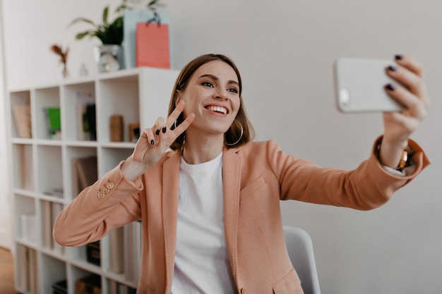 Portrait of smiling office worker with black manicure in soft pink jacket. Woman shows peace sign and makes selfie.