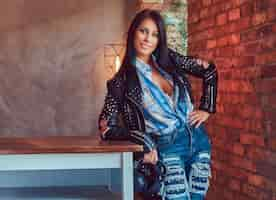 Free photo portrait of a smiling sexy sensual brunette posing in a stylish leather jacket and jeans leaning on a table in a studio with a loft interior.