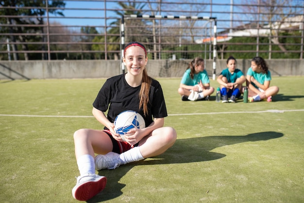 Portrait of smiling teenage football girl on field. Happy girl in sportswear resting on ground looking at camera while other girls sitting behind her enjoying talk. Active rest and team sport concept