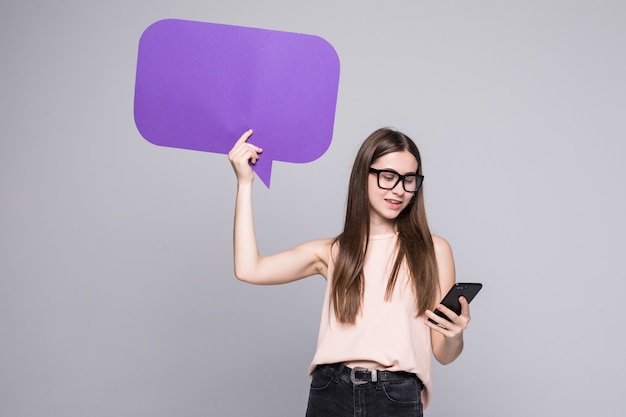 Portrait of a smiling woman showing blank speech bubble and holding mobile phone isolated over gray wall